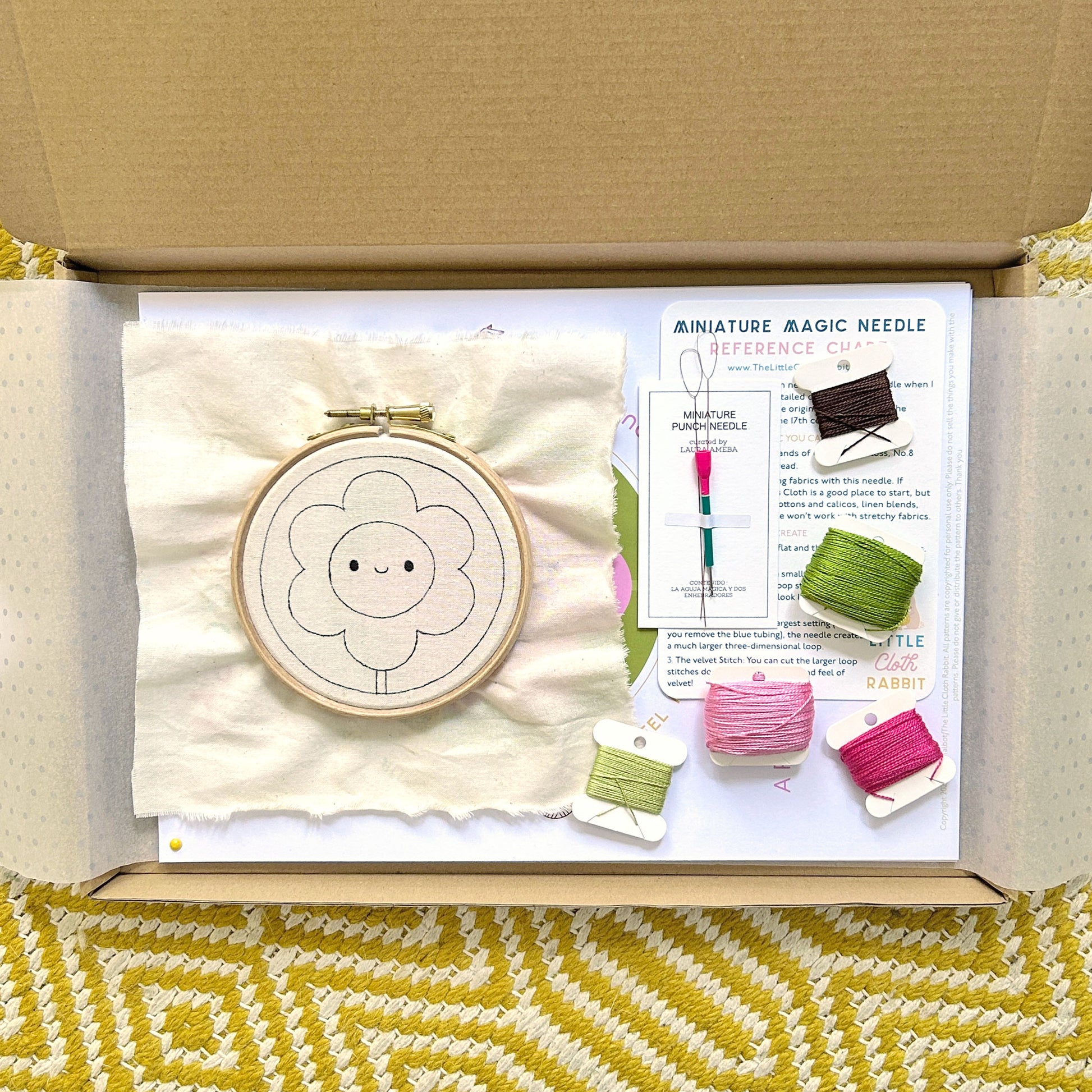 Opened punch needle kit box showing pre-drawn fabric in hoop, Miniature Magic Needle, and cotton threads.
