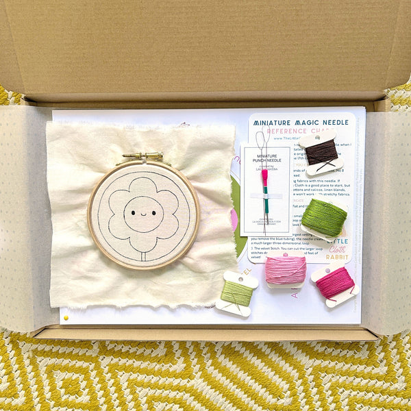 Opened punch needle kit box showing pre-drawn fabric in hoop, Miniature Magic Needle, and cotton threads.